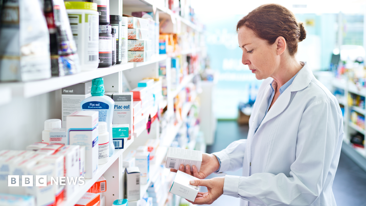 Pharmacist standing in front of shelves in pharmacy, holding medicine boxes in hands.
