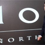 Andy Burnham walks past a black painted wall bearing the words No 10 North in white writing. He is wearing a dark suit, white shirt and dark blue tie and black-framed glasses. He has a serious expression on his face.