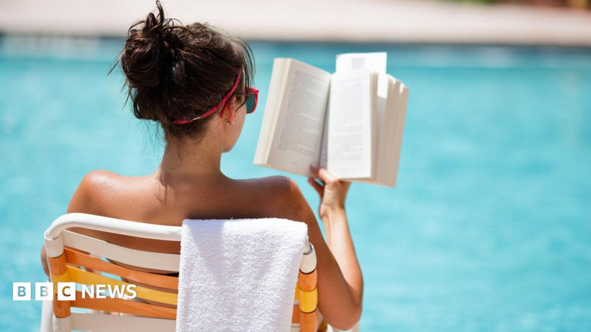 Young woman sitting in a chair with a towel draped over the back, reading at the pool in the summer