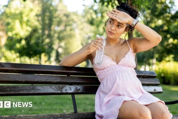 Pregnant woman sitting on a park bench in a light pink dress, holding a bottle of water.