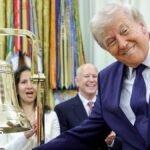 President Donald Trump ringing the opening bell in the Oval Office on Monday. The US president is smiling at the cameras as he rings the big gold bell. He is wearing a dark blue suite, with a dark blue spotted tie and a white shirt.