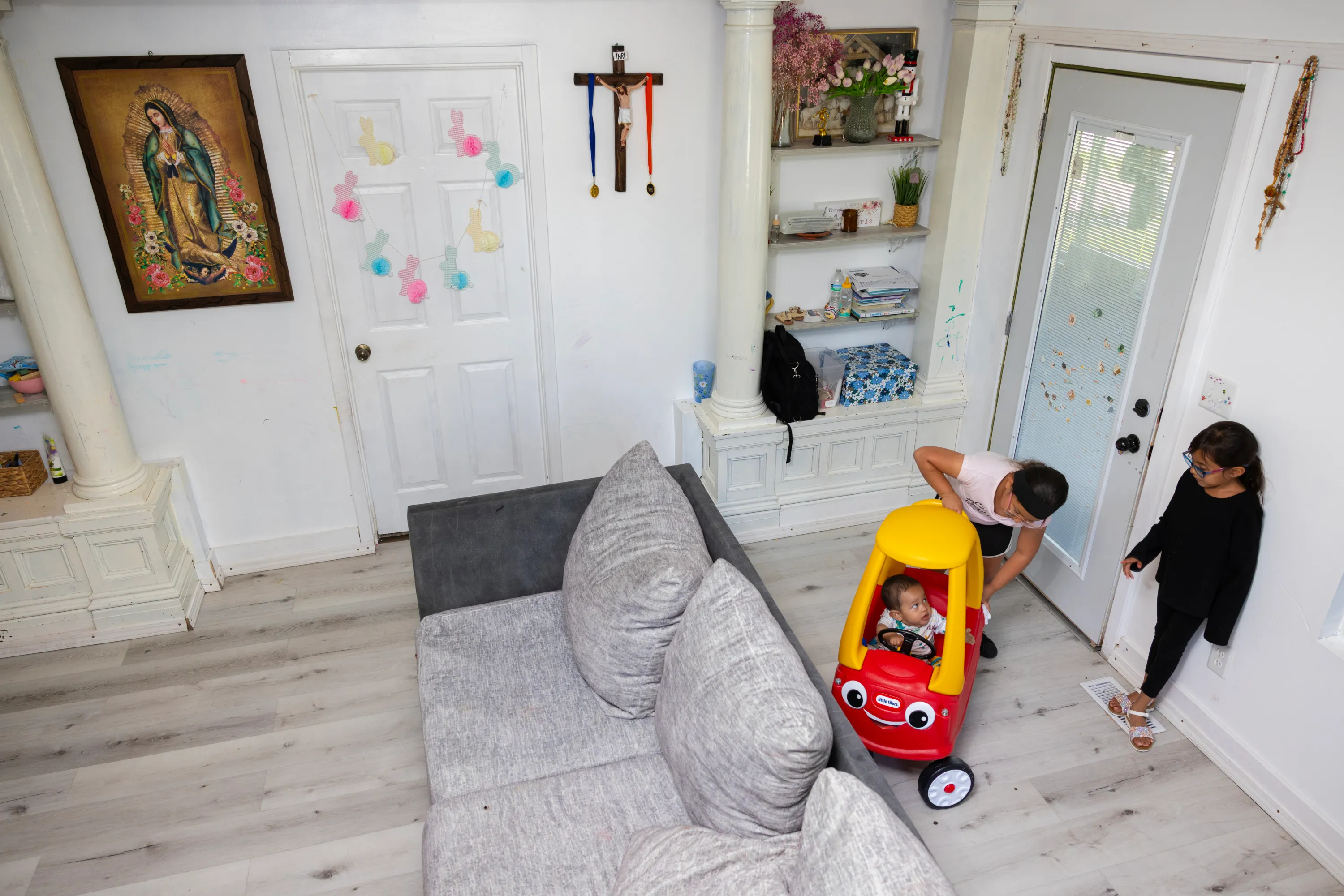 Two small children look at a baby in a yellow and red toy car inside a gray, tidy living room. There is a crucifix on the wall with red and blue medallions hanging from it. A painting of the Virgin of Guadalupe also decorates the wall.