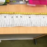 A long sheet of paper sits on wood-topped tables, with various boxes printed on it. A yellow tape measure is extended parallel to the paper.
