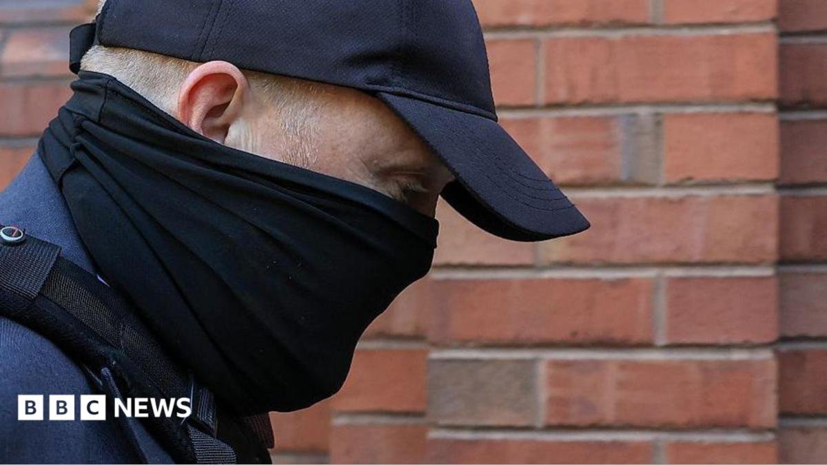 A close-up of the head and shoulders of a man with his face obscured by a black scarf and black cap as he walks towards a red-brick court. His eyes can be seen and his is looking downwards. He is wearing a blue suit jacket and the strap of a bag over his shoulder.