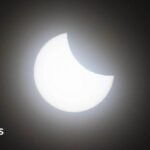 A picture of a partial eclipse showing the bright orange sun with a crescent over its lower half. The background is black.