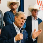 Texas Gov. Greg Abbott speaks into a microphone with his left hand raised in a gesture, addressing an audience. He is dressed in a navy suit jacket over a light-blue collared shirt.