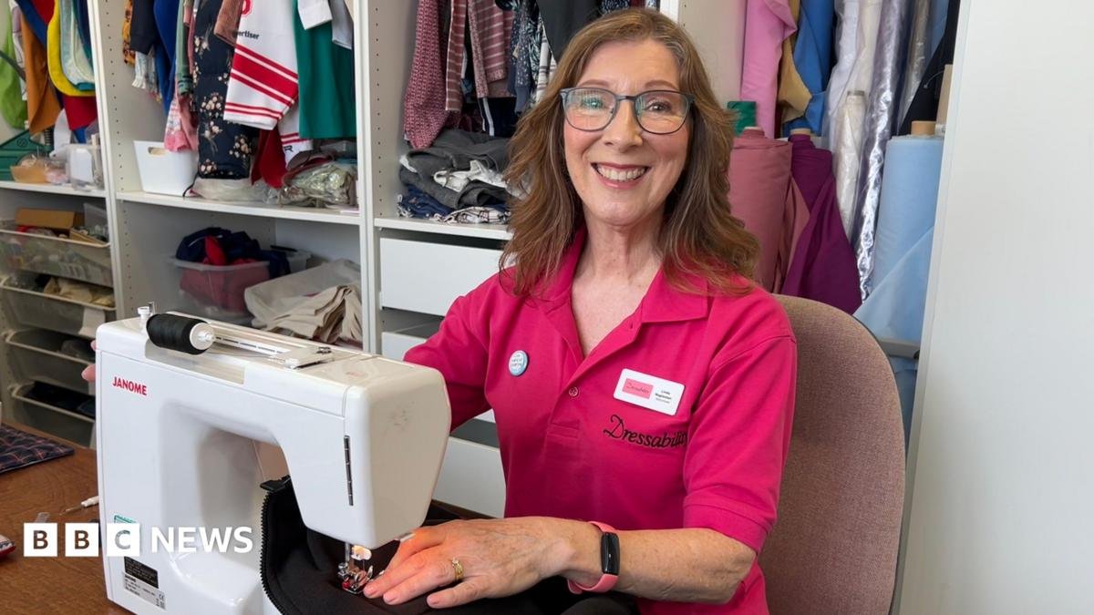 A woman with long brown hair and glasses is sitting at a sewing machine and smiling at the camera. She is wearing a pink collared shirt with the Dressability logo on it