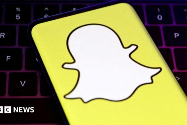 A closeup of Snapchat's logo, a white cartoon ghost with a bright yellow background, held over a laptop conputer keyboard.